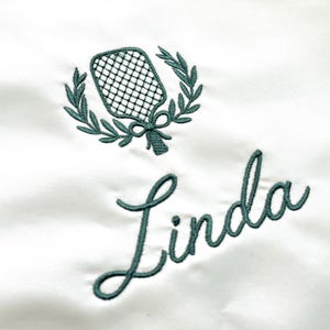 May include: A white fabric surface featuring dark green embroidery. The design includes a pickleball paddle centered within a laurel wreath, positioned above the name Linda written in a cursive script font using the same dark green thread.