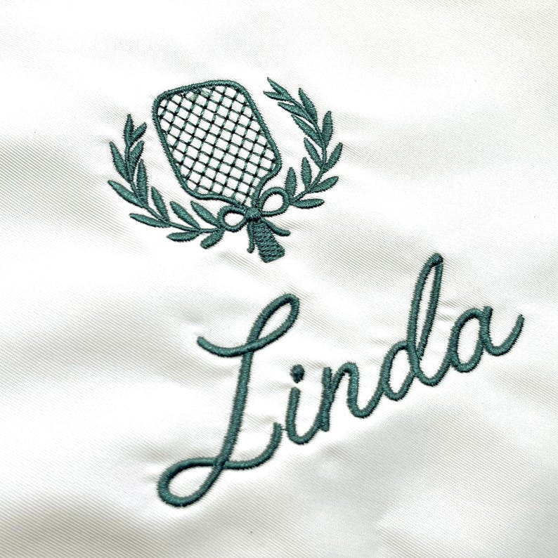 May include: A white fabric surface featuring dark green embroidery. The design includes a pickleball paddle centered within a laurel wreath, positioned above the name Linda written in a cursive script font using the same dark green thread.