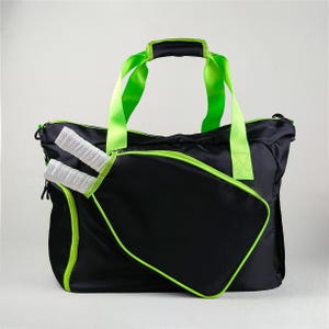 Op de afbeelding: Zwarte sporttas met neon groene accenten, waaronder de rits, handvatten en randen. De tas heeft een ritsvak aan de voorkant, met daarin twee sportrackets met witte en zwarte handvatten.