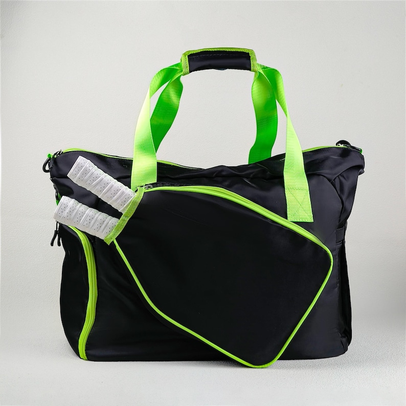 Op de afbeelding: Zwarte sporttas met neon groene accenten, waaronder de rits, handvatten en randen. De tas heeft een ritsvak aan de voorkant, met daarin twee sportrackets met witte en zwarte handvatten.