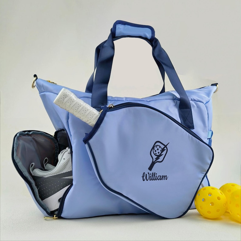 Op de afbeelding: Een lichtblauwe pickleballtas met marineblauwe accenten. De tas heeft een vak met ritssluiting voor schoenen en een vak voor een racket. Een racket is zichtbaar, samen met twee gele pickleballs. De naam "William" staat op de racketcover.