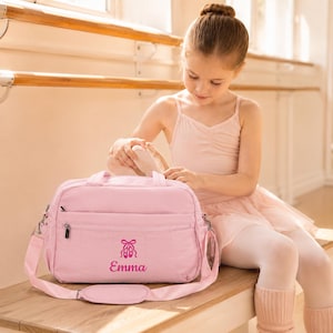 May include: A light pink rectangular duffel bag with a zippered front pocket, featuring a dark pink graphic of ballet slippers and the name Emma printed in a script font. The bag includes a shoulder strap with a padded section and silver hardware.