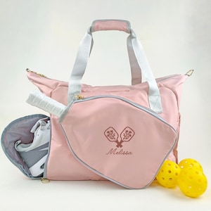 Op de afbeelding: Een roze pickleball-tas met een grijze rand, witte handvatten en een bijpassende pickleball-racketcase. De tas heeft een ritssluiting en een zijvak met een witte en grijze sportschoen. De tas heeft de naam "Melissa" en een pickleball-afbeelding.