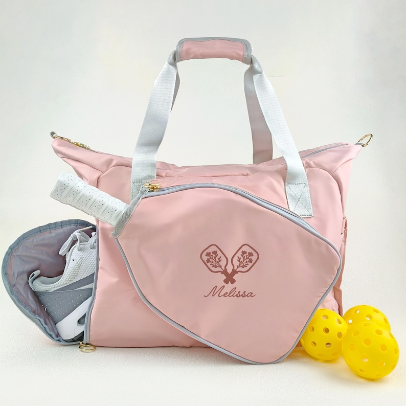 Op de afbeelding: Een roze pickleball-tas met een grijze rand, witte handvatten en een bijpassende pickleball-racketcase. De tas heeft een ritssluiting en een zijvak met een witte en grijze sportschoen. De tas heeft de naam "Melissa" en een pickleball-afbeelding.