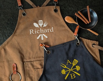 Puede incluir: Dos delantales con correas de cuero y herrajes metálicos. Uno es marrón con el nombre "Richard" y un gráfico de utensilios. El otro es azul marino con las letras "A" y "B" y un gráfico cruzado de tenedor, espátula y pesas. También se ven una cuchara de madera y palitos de canela.