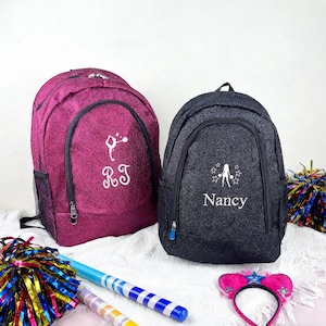Peut inclure: Deux sacs à dos scintillants, un magenta et un noir, sont présentés. Le sac à dos magenta porte les initiales "RJ" et un motif de gymnaste. Le sac à dos noir porte le nom "Nancy" et un motif d'étoile. Des accessoires de cheerleading sont également visibles.