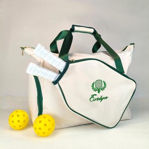 Op de afbeelding: Cr&egrave;mekleurige pickleballtas met groene accenten, handvatten en rits. Inclusief twee pickleball rackets met witte grips en twee gele pickleballs. De tas heeft de naam "Evelyn" in groen geborduurd.