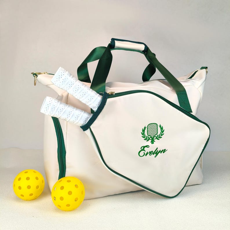 Op de afbeelding: Cr&egrave;mekleurige pickleballtas met groene accenten, handvatten en rits. Inclusief twee pickleball rackets met witte grips en twee gele pickleballs. De tas heeft de naam "Evelyn" in groen geborduurd.