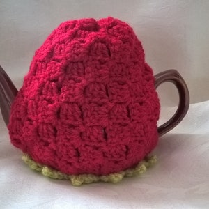 Op de afbeelding: Een rode, gehaakte theepotwarmer in de vorm van een aardbei. De warmer heeft een groene, gehaakte rand aan de onderkant.