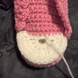 May include: Close-up of a pink and white crocheted slipper with a white yarn tail hanging down. The slipper is being sewn together.