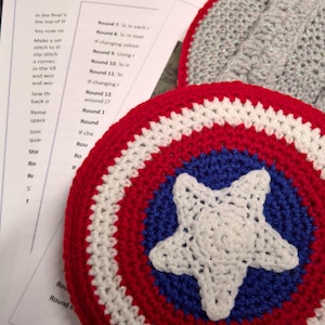 May include: A crocheted Captain America shield with red, white, and blue colors. The shield is made of yarn and has a star in the center.