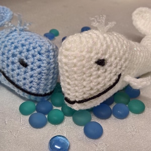 May include: Two crocheted whale toys, one blue and one white, with black eyes and mouths, are lying on a white surface with blue and green glass pebbles.
