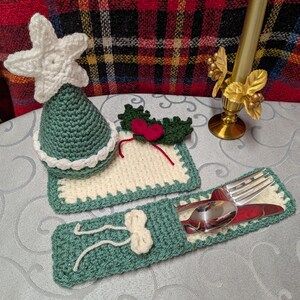 May include: Crocheted Christmas decorations including a tree topper, placemat, and cutlery holder. The tree topper is white and green, the placemat is cream with green trim, and the cutlery holder is green with a cream bow.