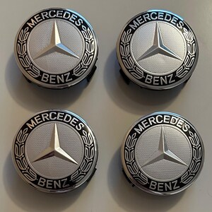 Op de afbeelding: Vier zilveren en zwarte Mercedes-Benz wieldopjes. Elke dop heeft het Mercedes-Benz-logo, een driepuntige ster, in het midden, omgeven door een laurierkransontwerp. Het woord "Mercedes" staat bovenaan en "Benz" onderaan.