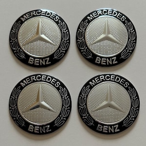 Puede incluir: Cuatro tapacubos Mercedes-Benz plateados y negros. Cada tapacubo presenta el logotipo de Mercedes-Benz, una estrella de tres puntas, en el centro, rodeado de un fondo plateado texturizado y un borde negro con las palabras "MERCEDES" y "BENZ".