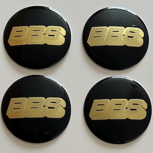 Könnte beinhalten: Vier schwarze, kreisförmige Radnabenabdeckungen mit den goldfarbenen Buchstaben "BBS" in fetter, serifenloser Schrift. Die Kappen haben eine glänzende Oberfläche und sind quadratisch angeordnet.