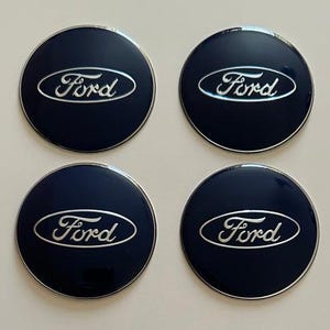 Puede incluir: Cuatro tapacubos Ford azul marino con el logotipo Ford plateado. Las tapas son redondas con un acabado brillante y un borde plateado. El logotipo de Ford está en cursiva.