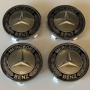 Può includere: Quattro coprimozzi Mercedes-Benz argento e neri. Ogni coprimozzo presenta il logo Mercedes-Benz, una stella a tre punte all'interno di un cerchio, e la scritta "Mercedes" sopra "Benz". I coprimozzi hanno una finitura metallica lucida.