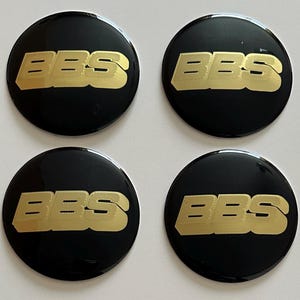 Może przedstawiać: Cztery czarne, okrągłe kołpaki BBS z złotymi napisami. Logo BBS jest wyświetlane złotą, teksturowaną czcionką na błyszczącym czarnym tle. Te kołpaki są prawdopodobnie przeznaczone do kół samochodowych.