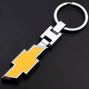 Puede incluir: Un llavero de metal plateado con un emblema de rayo amarillo. El emblema tiene un borde plateado y está unido a un cierre y un llavero de metal plateado. El llavero es probablemente para un coche o vehículo.