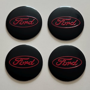 Puede incluir: Cuatro emblemas circulares negros con la palabra "Ford" en rojo en una escritura estilizada. Los emblemas son brillantes, probablemente de plástico o metal. El logotipo de Ford es prominente.