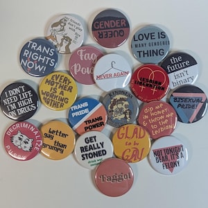 May include: A selection of circular badges with diverse slogans and designs. The badges display phrases such as "Trans Rights Now!", "Gender Queer", "Bisexual Pride!", and "Glad to be Gay". Colours include red, blue, yellow, and white.