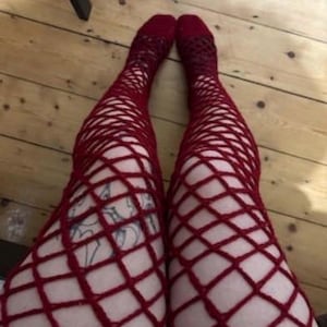 Puede incluir: Un par de medias de rejilla rojas. Las medias llegan hasta la rodilla y tienen un patrón en forma de diamante. Los calcetines son de color rojo intenso y se llevan sobre un suelo de madera.