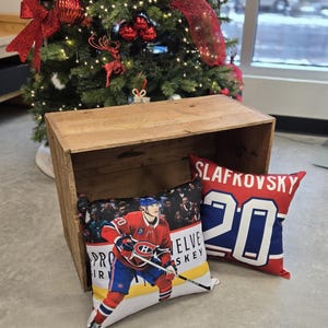 Könnte beinhalten: Zwei dekorative Kissen mit Hockey-Motiven. Ein Kissen zeigt ein rot-weiß-blaues Hockeytrikot mit der Nummer 20 und dem Namen "Slafkovsky" darauf gedruckt. Das andere Kissen zeigt ein Foto eines Hockeyspielers, der ein rot-weiß-blaues Trikot mit der Nummer 20 trägt. Der Spieler hält einen Hockeyschläger und befindet sich auf dem Eis. Das Kissen trägt die Worte "Pro Hockey" und "Elve Skey" aufgedruckt.