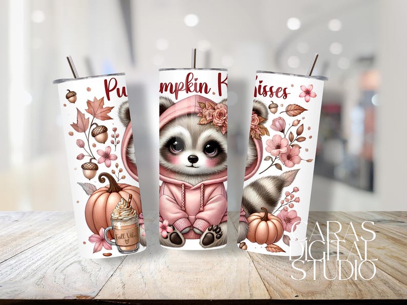 May include: White tumbler featuring a cute raccoon in a pink hoodie, surrounded by pumpkins, flowers, and fall leaves. The text "Pumpkin Kisses" is displayed on the tumbler. A fall-themed drink is also shown.