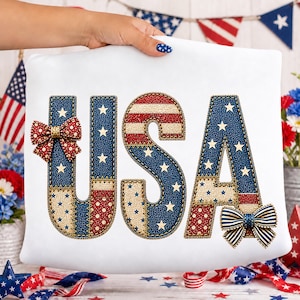 May include: White cushion featuring the word "USA" in a patchwork design. The letters are blue with white stars and red and white stripes. Decorative bows are attached to the letters. Patriotic theme.