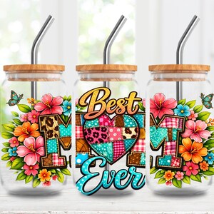 Op de afbeelding: Drie glazen bekers met houten deksels en metalen rietjes. Elke beker heeft een bloemenontwerp met het woord "MOM" en de tekst "Best Ever" op de middelste beker. Het ontwerp bevat een patchwork hart en vlinders.