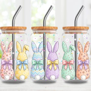 Puede incluir: Seis vasos de vidrio con pajitas de acero inoxidable y tapas de madera. Cada vaso presenta un diseño de conejito de Pascua colorido con un lazo. Los conejitos son de colores pastel, con detalles florales y estampados. El fondo tiene acentos florales y de lunares.