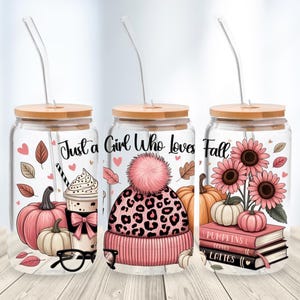May include: Three glass tumblers with wooden lids and clear straws. Each tumbler features a fall-themed design with pumpkins, leaves, and the text "Just a Girl Who loves Fall." One has a coffee drink, another a pink leopard print hat, and the last sunflowers.