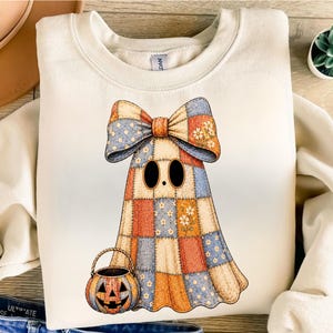 Puede incluir: Una sudadera de cuello redondo color crema con un gráfico de un fantasma hecho de tela de patchwork en tonos azul, naranja y blanco. El fantasma lleva un gran lazo de patchwork a juego y sostiene un pequeño cubo de dulces de calabaza naranja.