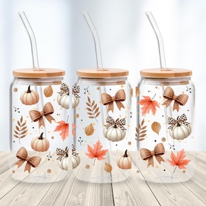 Puede incluir: Tres vasos de vidrio transparente con tapas de bambú y pajitas de vidrio curvadas. Cada vaso está decorado con ilustraciones temáticas de otoño, incluyendo calabazas, lazos y hojas en tonos marrones, naranjas y blancos. Los vasos están diseñados para bebidas frías.