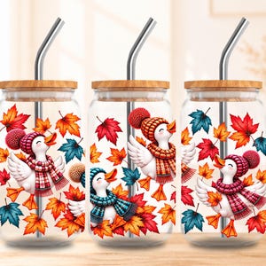 Puede incluir: Tres vasos de cristal transparente con tapas de bambú y pajitas de metal. Cada vaso presenta un estampado gráfico de patos blancos con gorros y bufandas de punto, rodeados de coloridas hojas de arce otoñales en tonos rojo, naranja y azul.