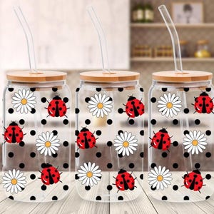 May include: Three clear glass drinking jars with bamboo lids and glass straws. Each jar is decorated with a pattern of red ladybirds, white daisies, and black polka dots. The jars are designed for cold beverages.