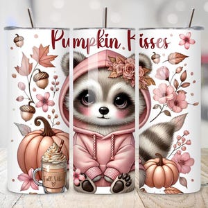 May include: A white tumbler with a fall theme. It features a raccoon wearing a pink hoodie, the text "Pumpkin Kisses", pumpkins, acorns, and floral accents. A coffee cup with the text "Fall Vibe" is also visible.