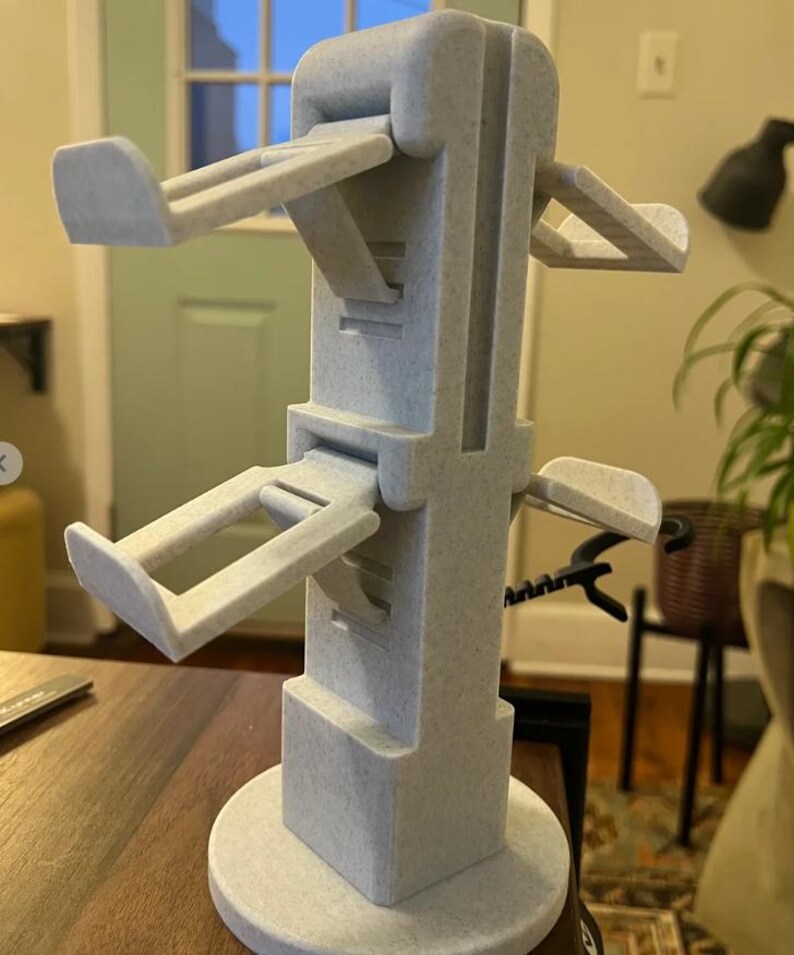 May include: A white 3D printed phone stand with four adjustable arms. The stand has a round base and a square column.