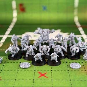 May include: A collection of gray miniature figures, possibly for a tabletop game, arranged on a green surface marked with white lines and a red "X". The figures vary in design, with some mounted on black bases and others are loose.