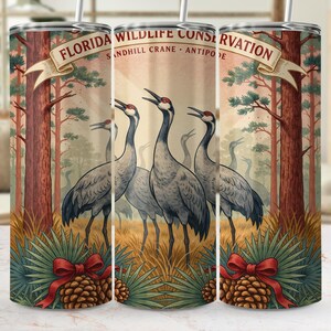 May include: A cylindrical tumbler featuring a graphic of Sandhill Cranes in a pine forest. The text reads FLORIDA WILDLIFE CONSERVATION, SANDHILL CRANE, ANTIPODE. The design includes pine trees, tall grass, pinecones, and red ribbons.