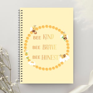Op de afbeelding: Een spiraalgebonden notitieboek met een lichtgele kaft. De kaft heeft een cirkelvormig ontwerp van honingraten en bijen, met de woorden "BEE KIND, BEE BRAVE, BEE HONEST" in een decoratief lettertype. Een vrolijk en inspirerend item.