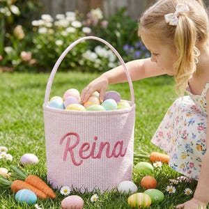 May include: A pink Easter basket filled with colorful eggs, with the name "Reina" embroidered on the side. The basket is on a grassy lawn, surrounded by more eggs and decorative carrots. A young girl is reaching into the basket.