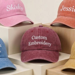 Könnte beinhalten: Fünf farbenfrohe Baseballkappen in Rosa, Blau, Rost, Burgunderrot und Gold. Jede Kappe hat eine individuelle Stickerei mit Namen oder Phrasen wie "Shirley", "Miranda Jones", "Jessie", "Custom Embroidery" und "THE PARTY".