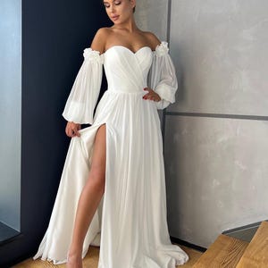May include: A white off-the-shoulder wedding dress with long, flowing sleeves and a thigh-high split. The dress has a sweetheart neckline and a fitted bodice.