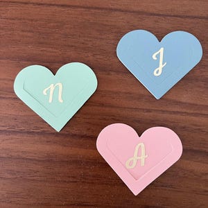 May include: Three heart-shaped paper cutouts in pastel colors: mint green, light blue, and pink. Each heart features a cursive letter in a contrasting gold color: "n", "J", and "A". The hearts are arranged on a dark wood surface.
