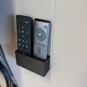 May include: A black rectangular remote control holder with two remotes. One remote is black, the other is silver. The holder is mounted on a white wall. The remotes have various buttons and symbols.