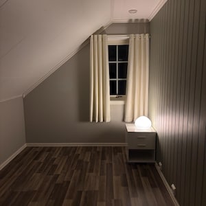 Puede incluir: Vista interior de un dormitorio con ventana, cortinas y mesita de noche. La habitación tiene techo inclinado y suelo de madera. Una lámpara esférica ilumina la mesita de noche. Las paredes están pintadas en tonos neutros.