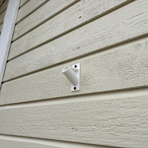 Puede incluir: Soporte de pared de plástico blanco con un brazo curvo, montado en un revestimiento de madera beige claro.