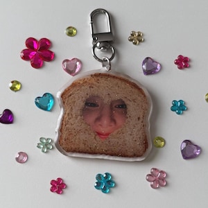 May include: A clear acrylic keyring featuring a slice of brown bread with a cutout in the centre revealing a human face. The keyring has a silver metal lobster clasp and is surrounded by small, colourful plastic gems shaped like hearts and flowers.